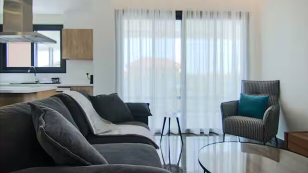 Een moderne woonkamer met een bank, een glazen salontafel en een stoel. Er is een keukenhoek met kasten en een raam met gordijnen.