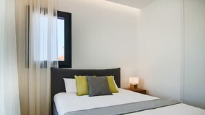 Ein ordentlich bezogenes Bett mit einer grauen Decke, einem dunklen Kopfteil und zwei Kissen in Grün und Grau. Ein Nachttisch mit Lampe und ein Fenster mit transparenten Vorhängen sind ebenfalls zu sehen.