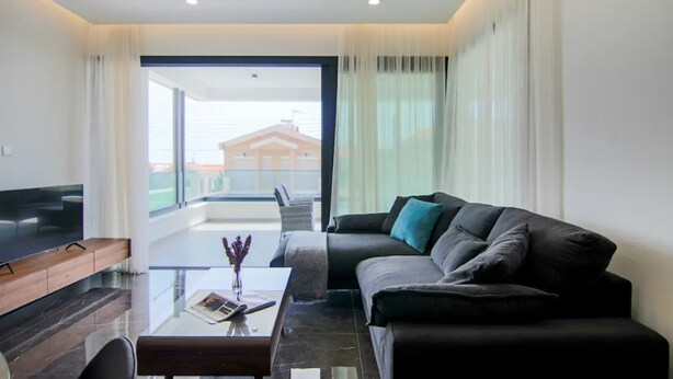 Ein modernes Wohnzimmer mit einem Flachbildfernseher, einem Glastisch, einer Couch mit Kissen und Blick auf ein Gebäude durch große Fenster.