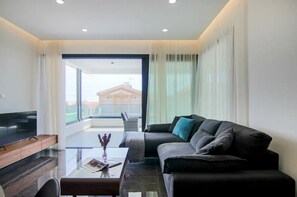 Un soggiorno moderno con una TV a schermo piatto, un tavolino in vetro, un divano con cuscini e vista su un edificio attraverso ampie finestre.