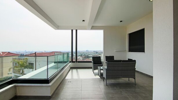 A modern balcony with a glass railing, a seating area with wicker furniture, and a city view. - 131 LIM 202 - Stay Anna Residence Sea View Apart (Limassol)