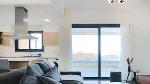 A modern living room with a sofa, a glass coffee table, and a view of a cityscape through a large window. - 131 LIM 202 - Stay Anna Residence Sea View Apart (Limassol)