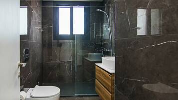 Un baño moderno con ducha de vidrio, inodoro blanco, tocador de madera y pared con azulejos de mármol.
