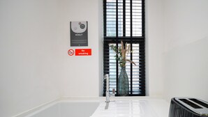 A modern bathroom with a large white bathtub, a no-smoking sign, a window with blinds, and a vase with dried plants. - Stay Awhile - Boutique Studio Northern Quarter (Manchester)
