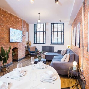A modern living room with a brick wall, a sofa, a dining table set for four, and a fireplace. - Stay Awhile | Stylish Industrial 2BR Duplex, NQ (Manchester)