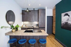 A modern kitchen with dark wood cabinets, a marble countertop, and a central island with blue bar stools. - Stay Awhile - Industrial 1BR Flat Northern Quarter (Manchester)
