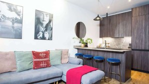 Ein modernes Wohnzimmer mit grauem Sofa, roten und grünen Kissen, einer roten Decke sowie einer Küchenzeile mit Bar und Hockern.