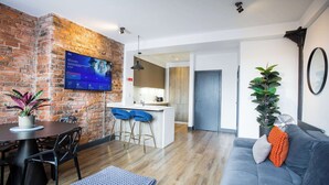 A modern living room with a brick accent wall, a flat-screen TV mounted above a dining table, a grey sofa, and a kitchen area in the background. - Stay Awhile - Luxury 2BR Duplex Northern Quarter (Manchester)