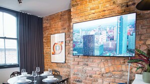 A dining area with a brick wall, a cityscape painting, and a modern table setup. - Stay Awhile - Luxury 2BR Duplex Northern Quarter (Manchester)