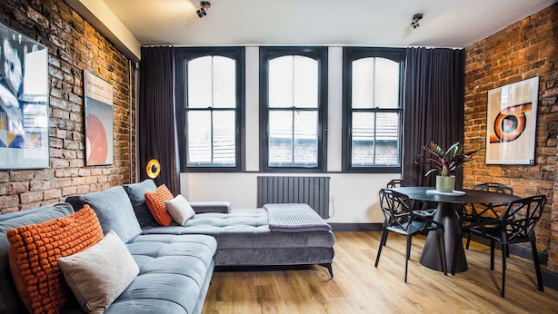 A modern living room with a grey sectional sofa, a round dining table with black chairs, large windows with dark curtains, and a brick wall with artwork. - Stay Awhile - Luxury 2BR Duplex Northern Quarter (Manchester)