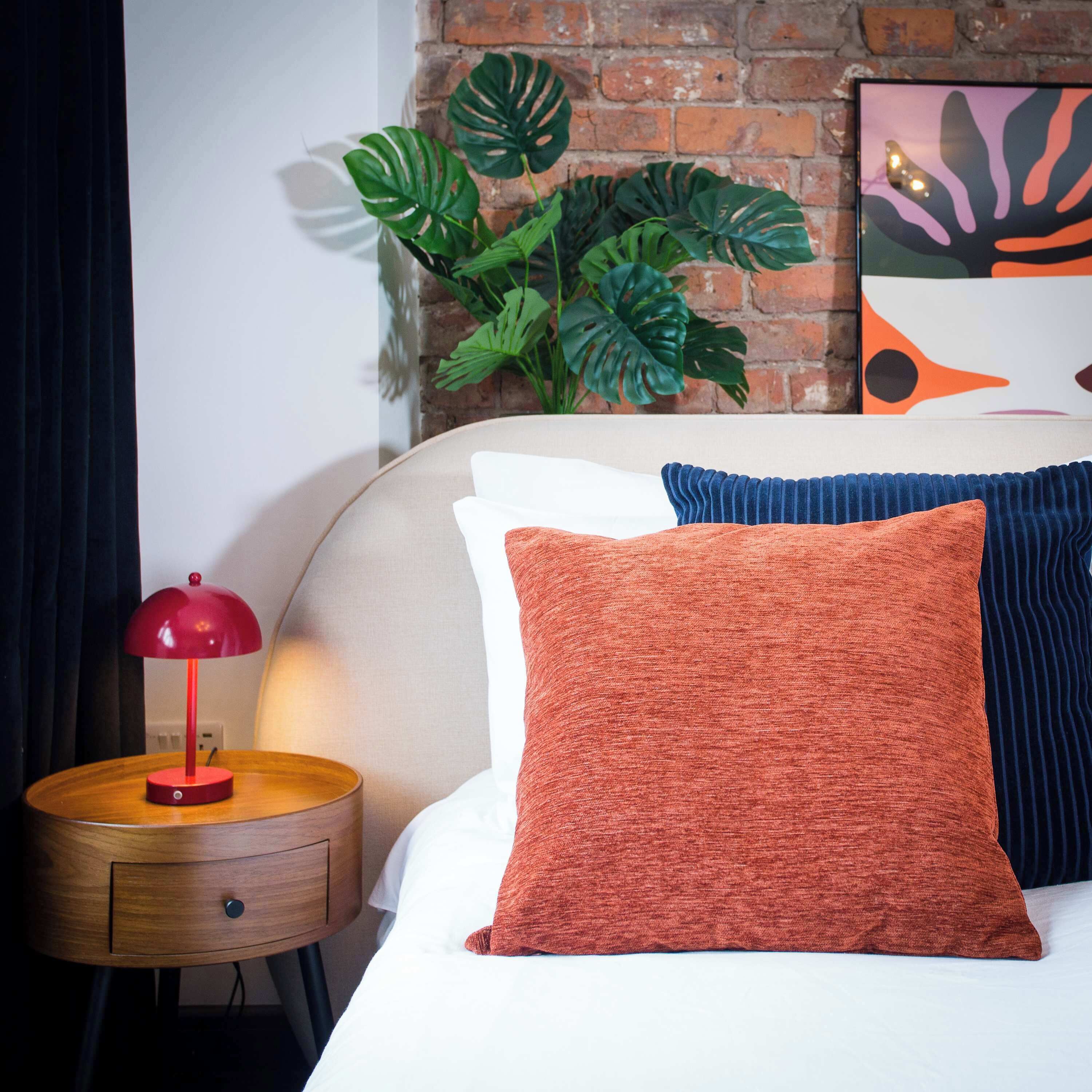A neatly made bed with two pillows, a small bedside table with a lamp, and a plant against a brick wall.