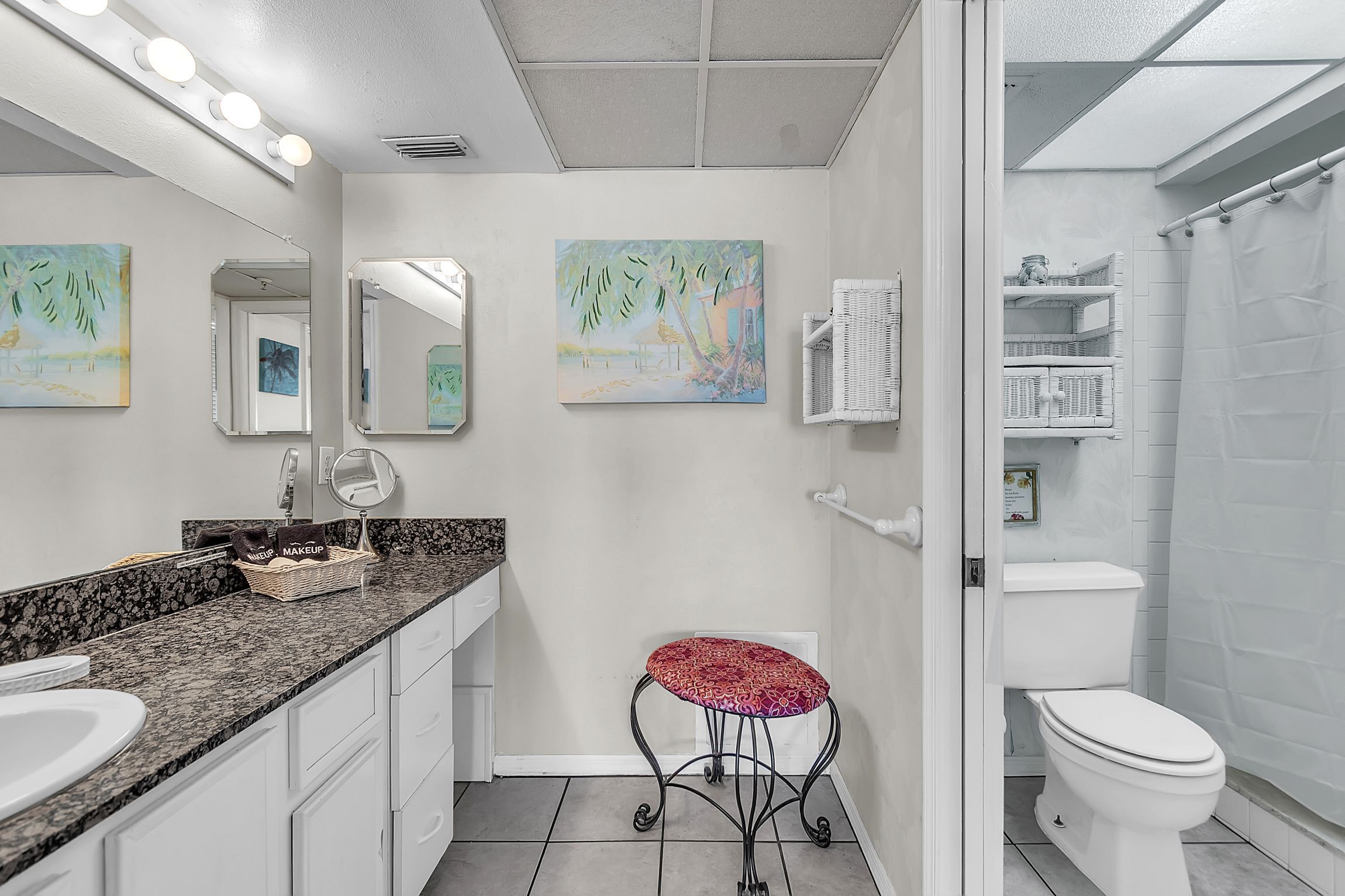 A bathroom with a white sink, vanity, and a toilet. There are two wall-mounted mirrors, a red stool, and a shower curtain.