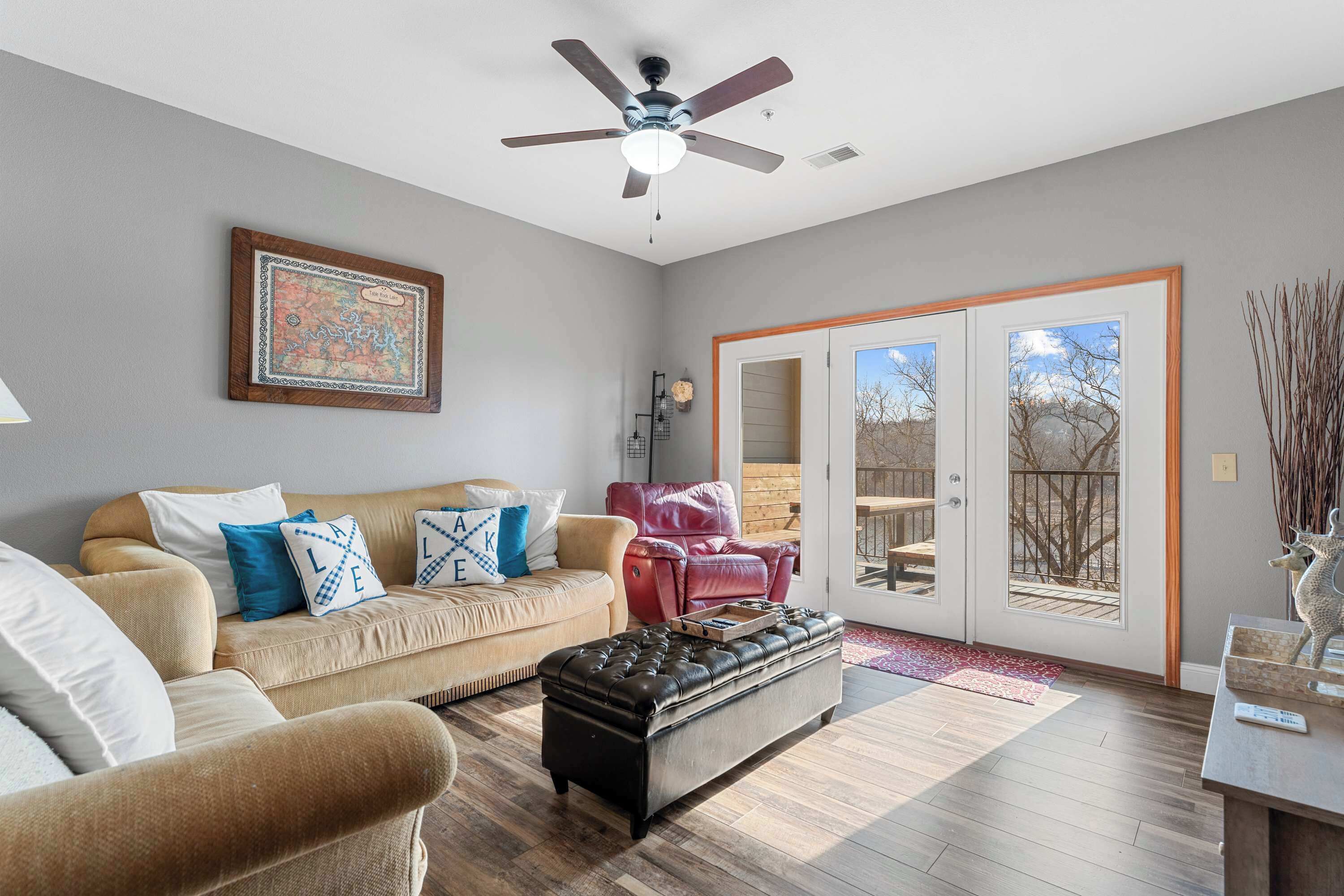 A living room with a beige sofa, a black ottoman, a ceiling fan, and a sliding glass door leading to an outdoor deck.
