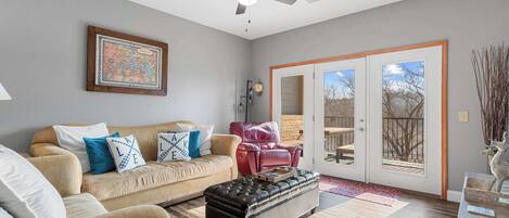 A living room with a beige sofa, a black ottoman, a ceiling fan, and a sliding glass door leading to an outdoor deck.