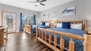 Ein Hotelzimmer mit zwei Betten, Holzkopfteilen, einem Deckenventilator, einem großen Fenster mit blauen Vorhängen und einem Holztisch.
