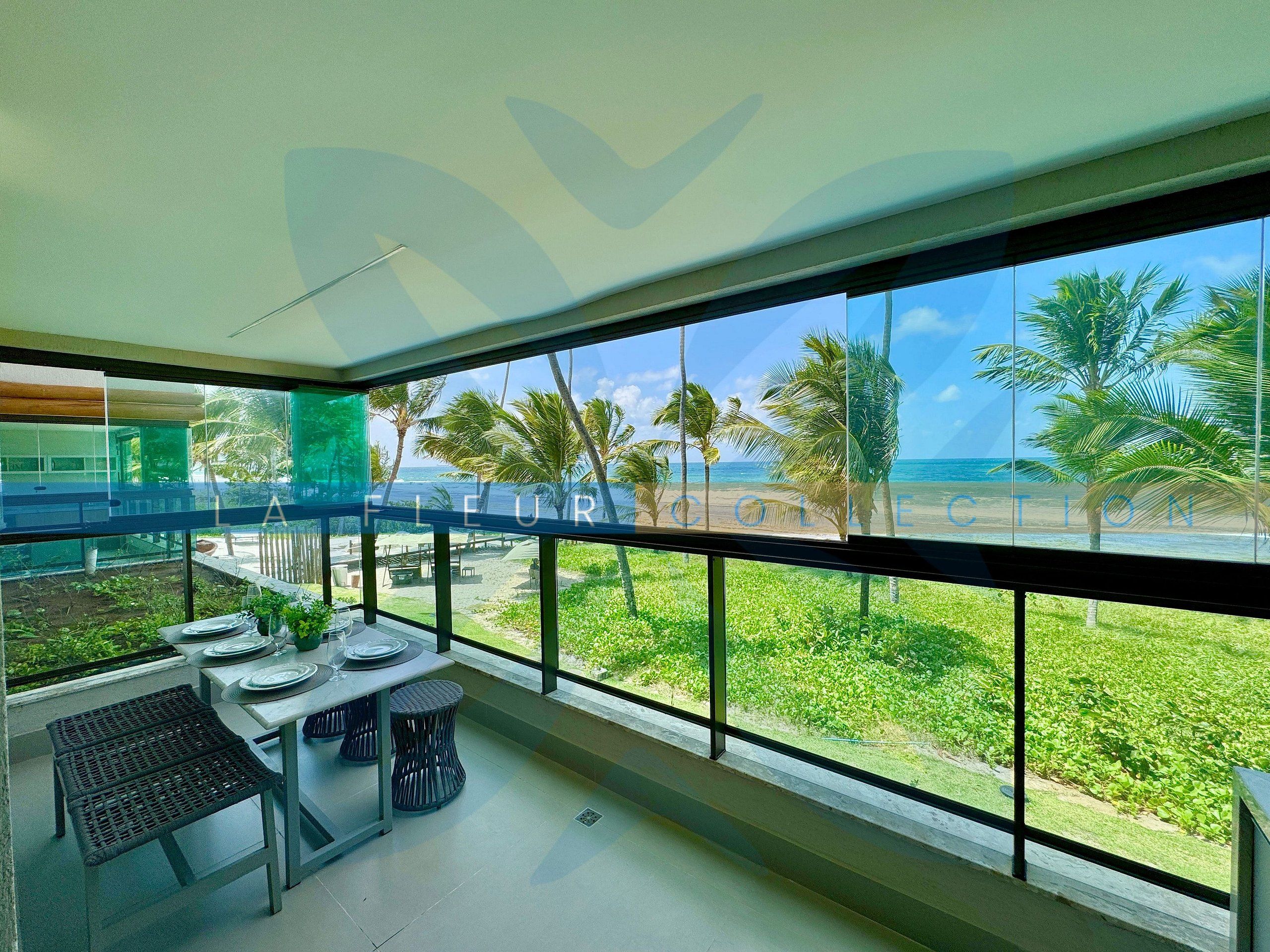 Varanda com mesa e cadeiras, vista para uma praia com palmeiras e o oceano.