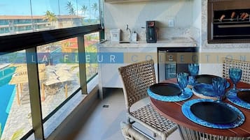 Uma sala de jantar com cadeiras de vime e uma mesa posta para dois, com vista para uma piscina e palmeiras.
