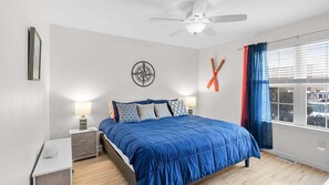 Ein Schlafzimmer mit einem Bett, zwei Nachttischen, einem Deckenventilator, einem Fenster mit Vorhängen und einer Wanddekoration.