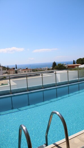 A clear blue pool with metal railings, a view of the sea, and buildings in the background. - 147 PEY - Stay Aloha Escape Apartment (Pegeia)