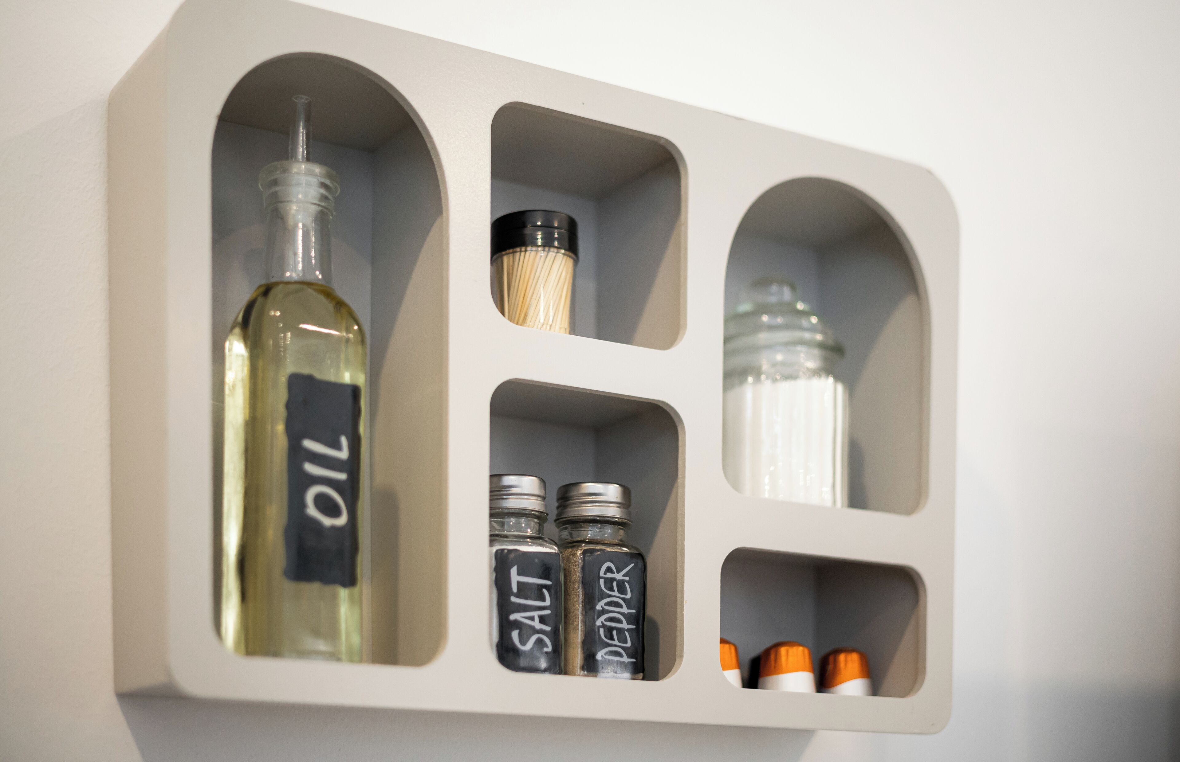 Eine wandmontierte Aufbewahrungseinheit mit Fächern, die eine mit "OIL" beschriftete Flasche, zwei mit "SALT" bzw. "PEPPER" beschriftete Gewürzbehälter und zwei kleine Behälter mit orangen Verschlusskappen enthält.