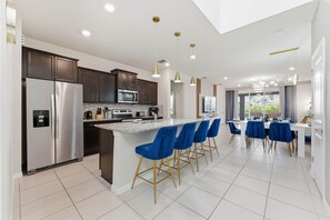 Une cuisine moderne dotée d’appareils électroménagers en acier inoxydable, d’un îlot central avec des tabourets de bar et d’un coin repas comprenant une grande table et des chaises bleues.
