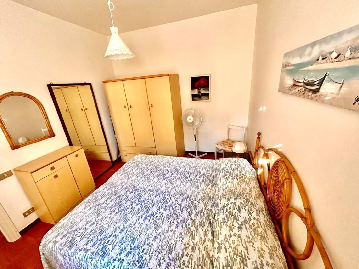 Een slaapkamer met een bed, een kledingkast, een spiegel, een ventilator en een schilderij aan de muur.
