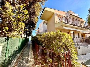A narrow pathway flanked by a green fence and a metal fence leading to a two-story building with balconies. - Three-room apartment with sea and beach view (Marcelli)