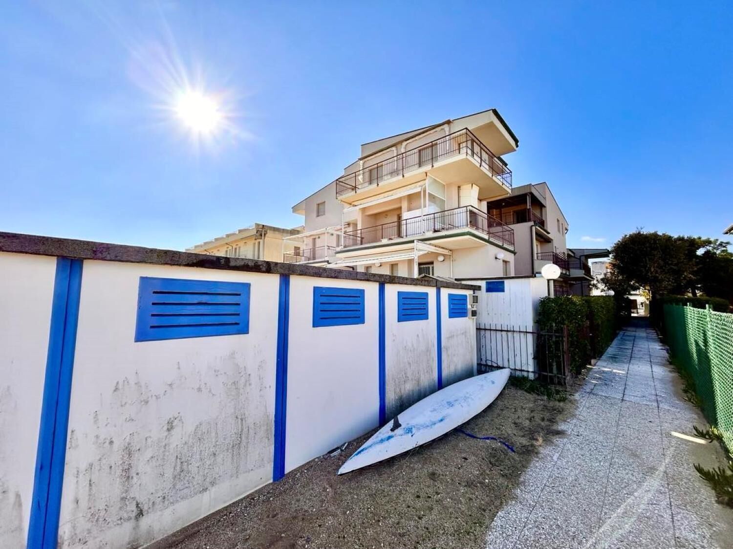Un edificio costero con varios balcones, una pared blanca con detalles azules y una tabla de surf en el suelo.