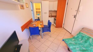 Ein gemütliches Zimmer mit Esstisch, zwei Stühlen, Fernseher, Bett mit gemustertem Bettbezug und einer Küchenzeile inklusive Waschmaschine.