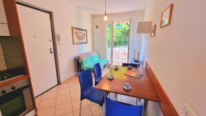 A compact kitchen with a stove and oven, a dining area with a table and blue chairs, and a view of a balcony with outdoor furniture. - Two-room Caesar [center, park, terrace] (Sirolo)