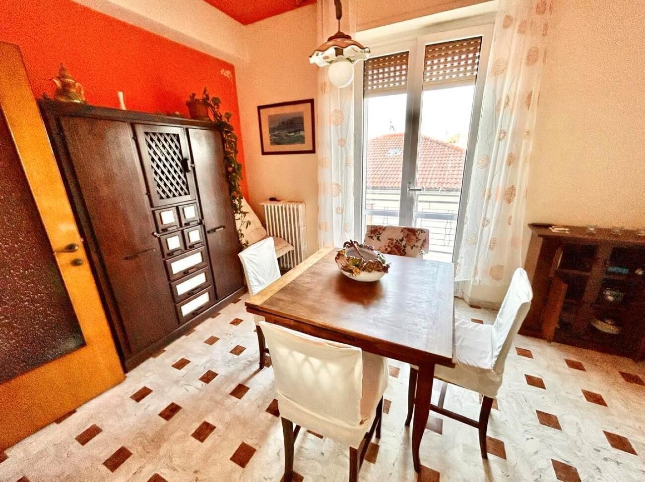 A dining area with a wooden table and chairs, a radiator, and a window with a view of buildings.