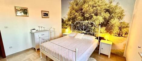 Un dormitorio con cama, mesitas de noche, lámpara de pared y un ventanal con vistas a árboles.