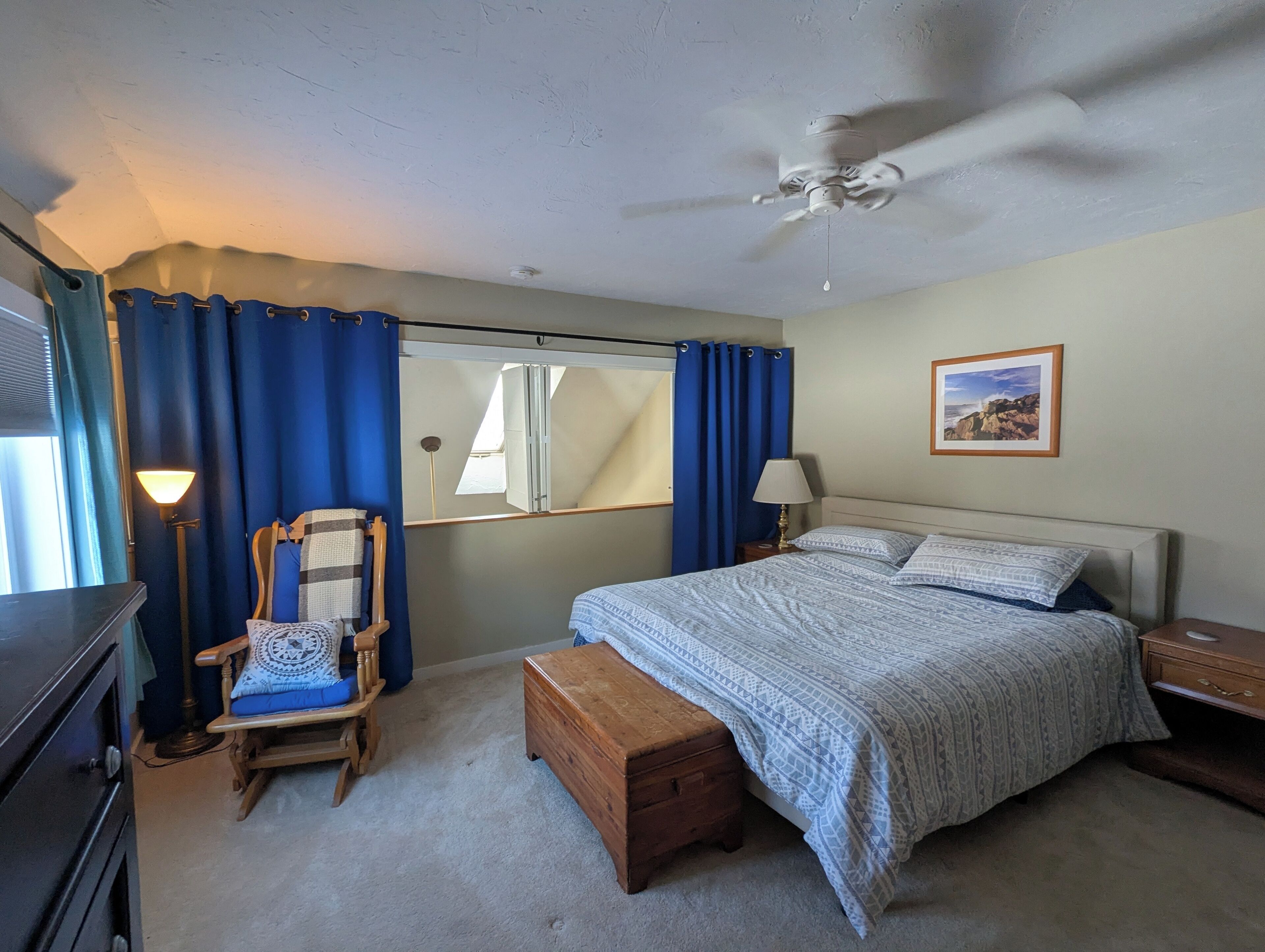 Une chambre à coucher comprenant un lit, un banc en bois, une chaise, un bureau, une lampe, un ventilateur de plafond et un tableau encadré accroché au mur.