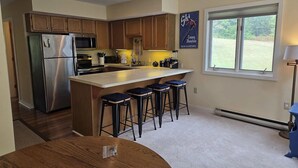 A modern kitchen with wooden cabinets, a stainless steel refrigerator, a central island with bar stools, and a window with a view of greenery. - Tenney Mountain Ski Resort Condo (Plymouth)