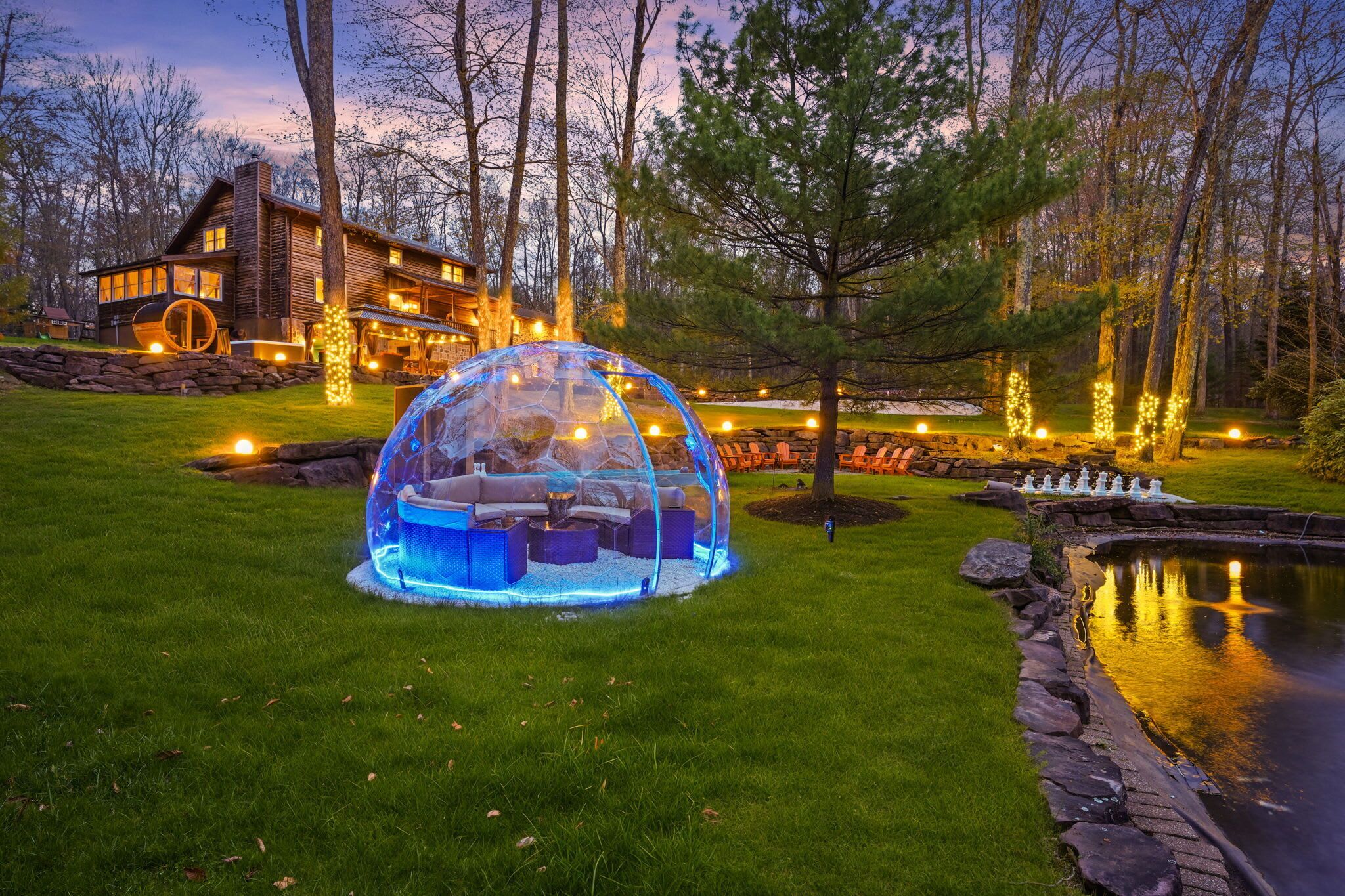 Un igloo de verre doté de sièges et d’un foyer, entouré d’un mur de pierre et d’un bassin, avec une maison en bois en arrière-plan.