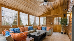 A cozy log cabin interior with a wooden ceiling, large windows with sheer curtains, a central seating area with sofas and cushions, and a door with a plant beside it. - Luxury meets Wilderness in the Heart of Poconos (Albrightsville)