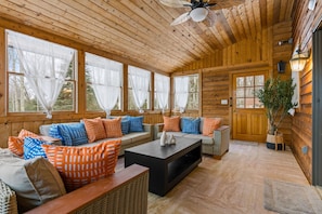 Ein gemütliches Blockhaus-Interieur mit Holzbalkendecke, großen Fenstern mit transparenten Vorhängen, einer zentralen Sitzecke mit Sofas und Kissen sowie einer Tür mit Pflanze daneben.