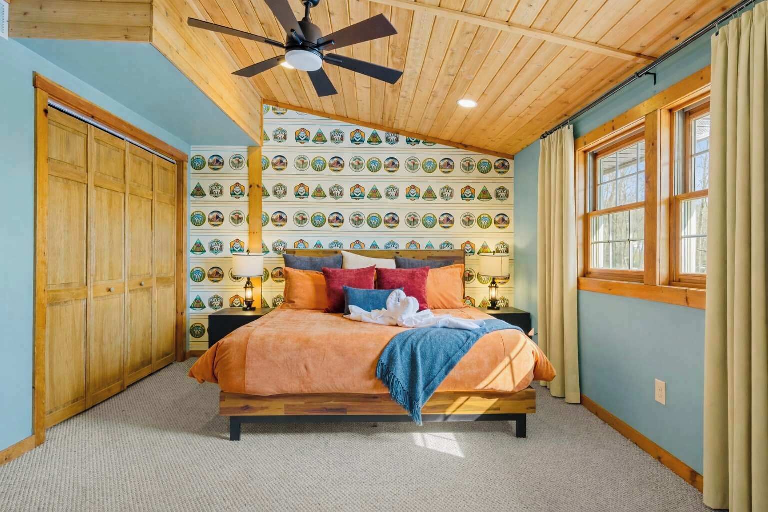 A bedroom with a wooden ceiling fan, a bed with colorful bedding, a wall with circular patterns, and a window with curtains.