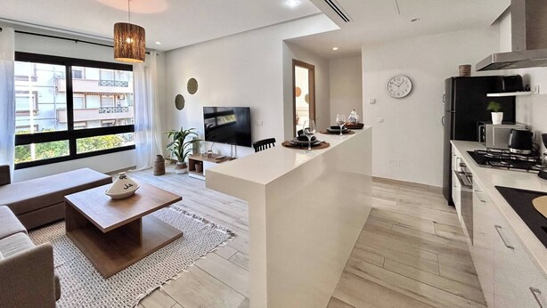 A modern kitchen with a central island, a dining area with a sofa and coffee table, and a large window with a view of buildings. - Malachite Elegance & Comfort in Sidi Daoud (La Marsa)