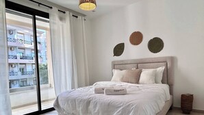 A bedroom with a bed, a view of buildings through a large window, and a woven rug on the floor. - Malachite Elegance & Comfort in Sidi Daoud (La Marsa)