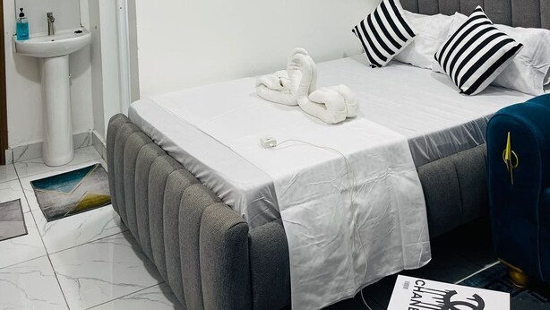 Una camera da letto moderna con un letto imbottito grigio, biancheria da letto bianca e cuscini a righe. Un piccolo vaso bianco con piante essiccate è appeso al muro.