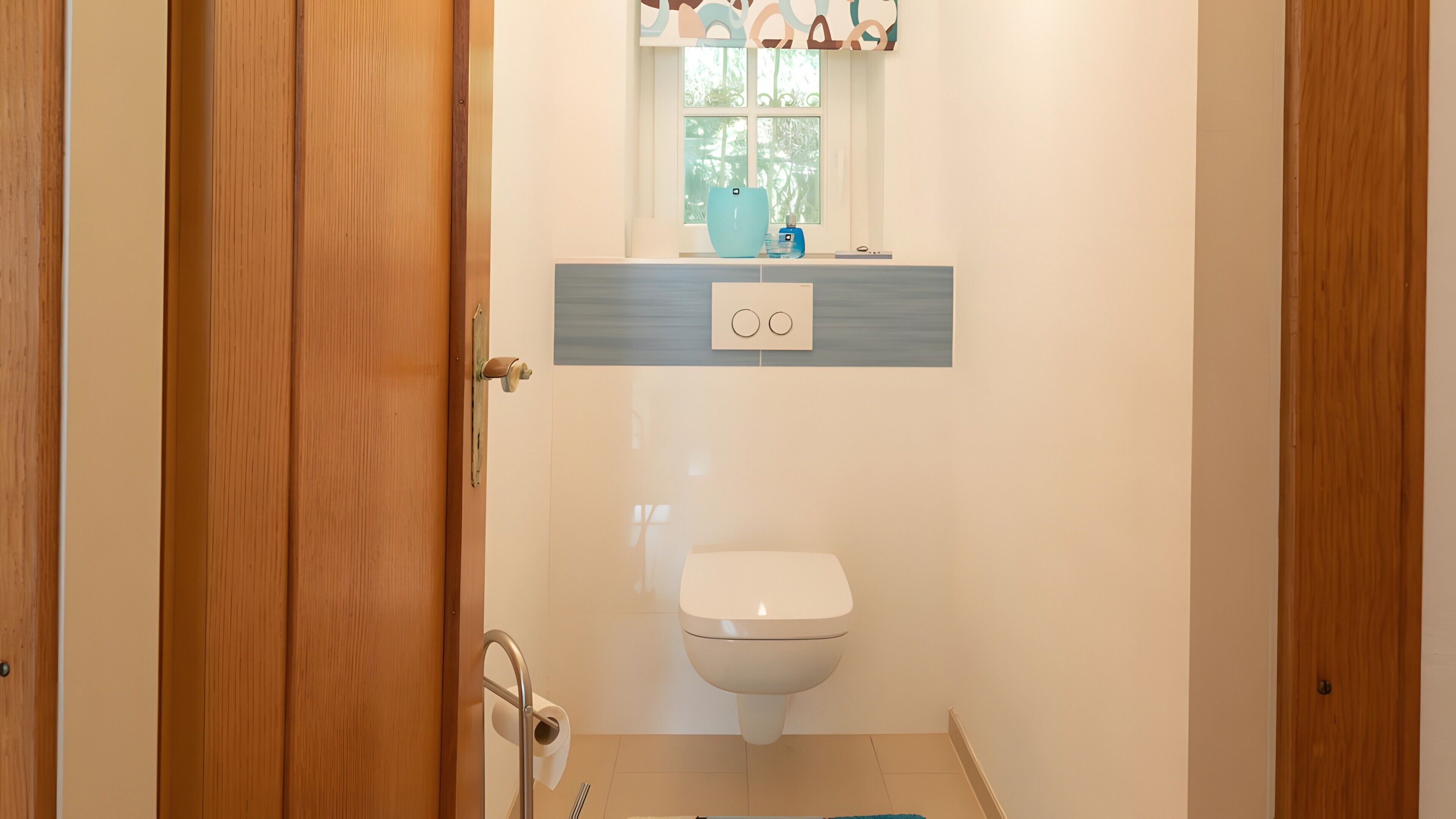 A bathroom with a toilet, a shelf with a blue vase, and a window with greenery outside.