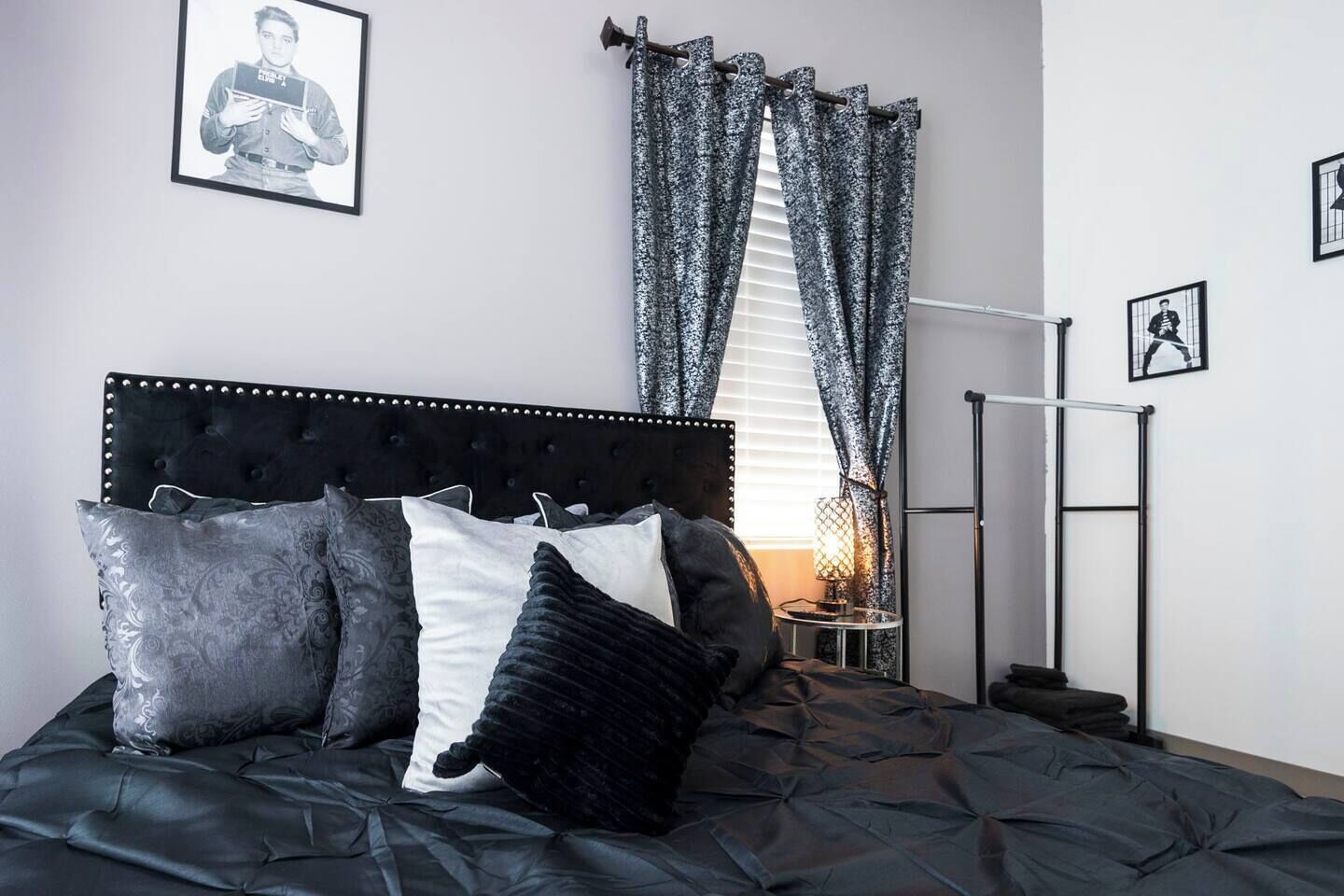 Une chambre à coucher avec une tête de lit capitonnée, un lit agrémenté d’oreillers à motifs, une table de chevet avec une lampe et une fenêtre avec des rideaux.