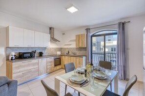 A modern kitchen with wooden cabinets, a built-in microwave, and a dining area with a table set for two. - Modern and cosy 2 bedroom flat with balcony BSAI1-1 (Swieqi)