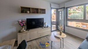 A modern living room with a TV on a wooden console, a small round table with a vase, and a balcony with a view of buildings and greenery. - Chic 2 bedroom Apartment with Comunal Pool VM75 (Villamartín)