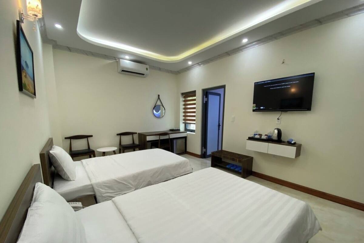 A hotel room with two beds, a desk, a chair, a TV, and a small shelf with items.