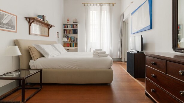 Un dormitorio con cama, mesita de noche, cómoda, estantería y un espejo grande.