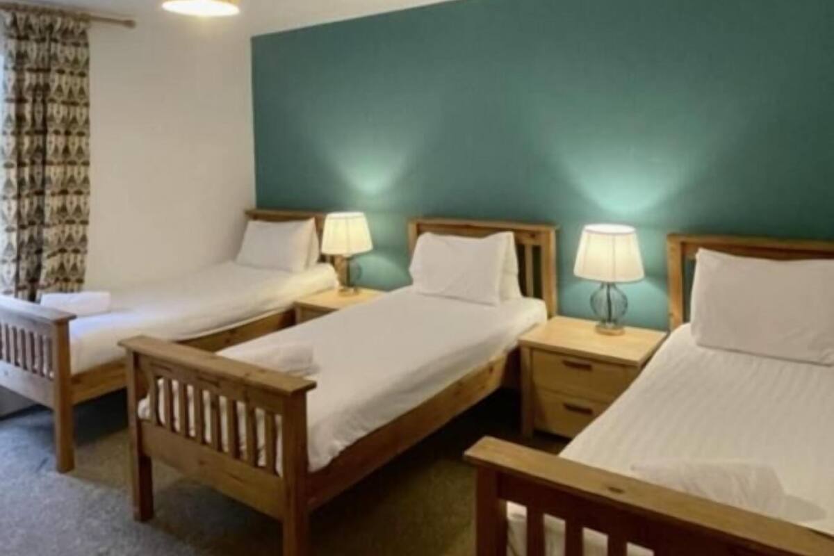 A room with three wooden beds, each with a nightstand and a lamp. The room has a teal accent wall and a ceiling light fixture.