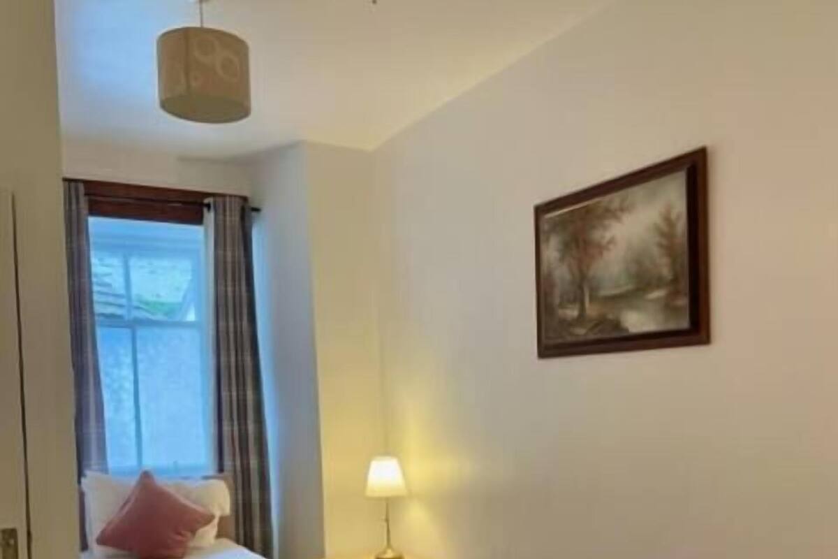A hotel room with two beds, a window with curtains, a painting on the wall, a lamp on a bedside table, and a ceiling light.