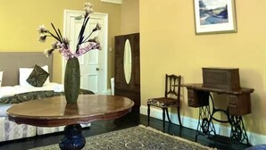 Une pièce avec un lustre, une table ronde ornée d’un vase de fleurs, un lit avec une tête de lit, un tableau encadré au mur et une machine à coudre.
